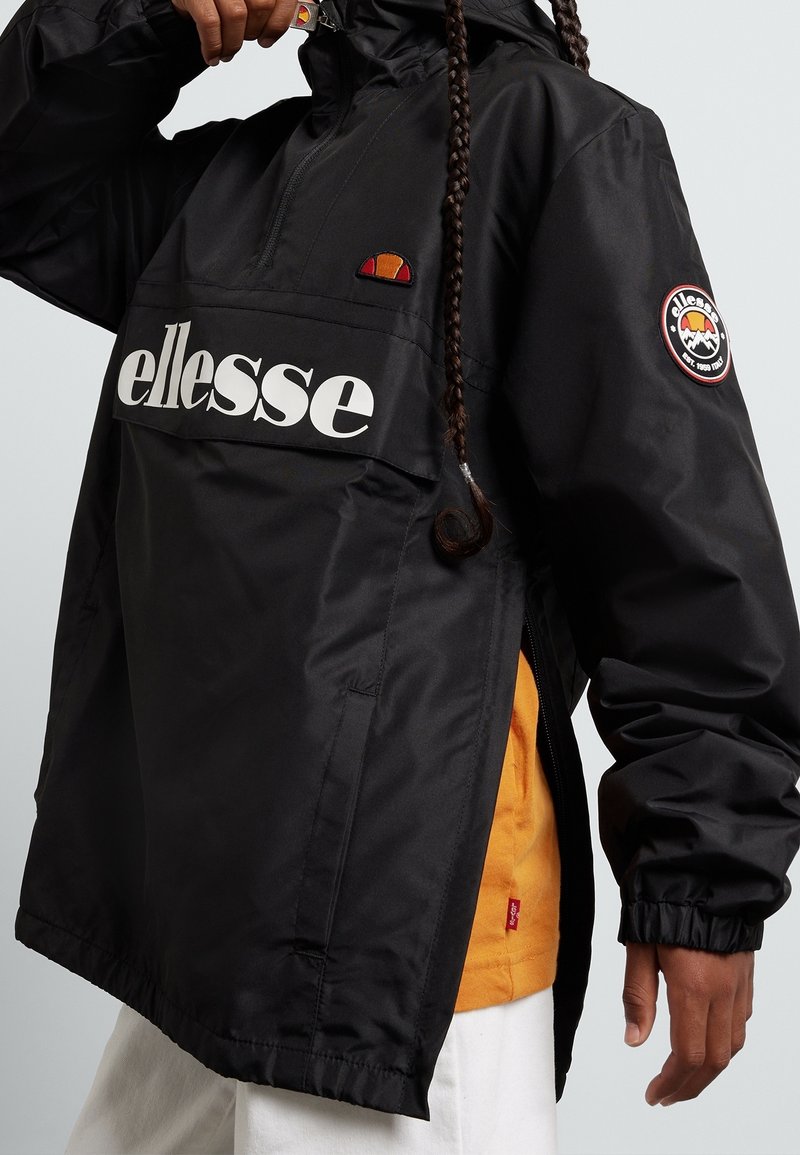 Personne portant une veste noire ellesse avec un logo sur la poitrine et la manche, dézippant partiellement le col montant, montrant un vêtement orange en dessous.