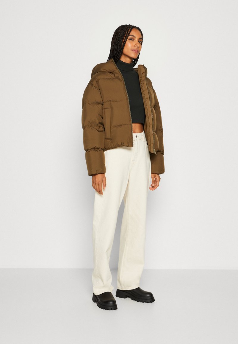 Braune, kurz geschnittene Pufferjacke mit Kapuze, Reißverschluss und gestepptem Obermaterial, kombiniert mit weißen, weiten Hosen und schwarzen, klobigen Stiefeln.