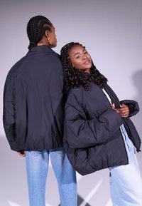 Casacos de penas pretos com designs oversized, apresentando textura acolchoada e bainhas elásticas. Os modelos usam jeans de ganga azul claro.