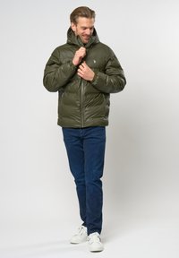 Alyvuogės spalvos pūkinė striukė su aukštu apykakle, užtrauktuku priekyje ir elastingomis rankovėmis. Derinama su tamsiai mėlynomis džinsomis ir baltomis sportinėmis bateliais.