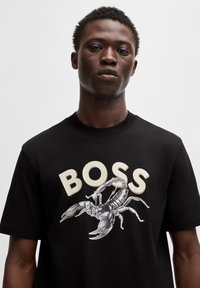Juodas medvilnės marškinėliai su dideliu skorpiono grafika ir žodžiu "BOSS" kreminės spalvos rašmenimis priekyje.