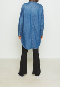 Camicia di jeans con maniche lunghe e polsini abbottonati, caratterizzata da un orlo arrotondato e dettagli sul girocollo posteriore, abbinata a pantaloni neri a zampa e sneakers.