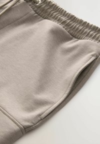 Pantalon de survêtement gris clair avec une texture douce, une taille élastique et des poches latérales. Le tissu semble décontracté et confortable.