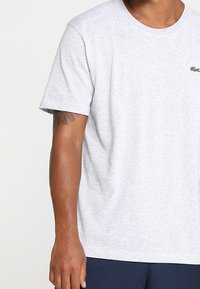 Šedé bavlněné tričko s krátkými rukávy, s kulatým výstřihem a malým zeleným logem na levé straně hrudi. Hladká textura.