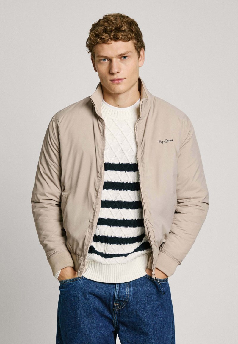 Beige vadderad jacka med dragkedja och ståkrage, bärs över en vit tröja med marinblå ränder och diamantmönster, kombinerad med blåjeans.