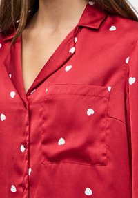 Blouse en satin rouge avec des motifs de cœurs blancs, une poche poitrine et un col, portée par une personne avec de longs cheveux bruns.