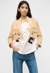 Γυναίκα με μακριά σκούρα μαλλιά φοράει μπεζ cropped σακάκι πάνω από λευκό πουκάμισο με μαύρο σχέδιο, συνδυασμένο με μπλε τζιν, τα χέρια στις τσέπες.