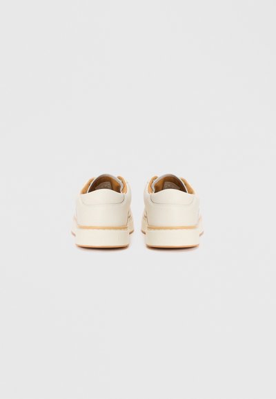 Sneakers brancos de pele com detalhes laranja, apresentando um acabamento suave, forma arredondada e uma sola de borracha texturizada. Marca no calcanhar.