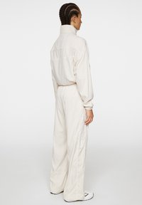 Ljus beige jumpsuit med hög krage, elastisk midja och vida ben. Har sydda detaljer och märkning längs sidan.