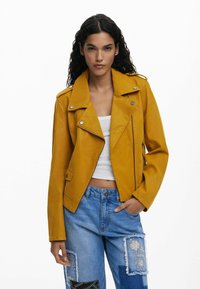 Gelbe Leder-Bikerjacke mit eingekerbtem Kragen, Reißverschlusstaschen und Druckknöpfen, getragen über einem weißen Oberteil und einer zerrissenen Jeans.