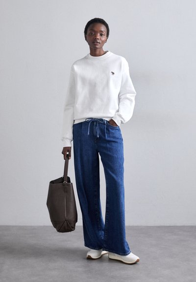 Hanorac alb cu un grafic mic, pantaloni din denim bleu cu croială largă, pantofi bej și o geantă tote maro închis. Texturi netede și croială casuală.
