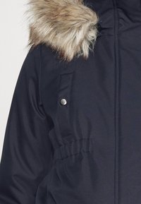 Laivastonsininen parkatakki, jossa tekoturkiskapussi, etutasku nepparikiinnityksellä sekä joustava vyötärösauma varmistamassa istuvuuden.