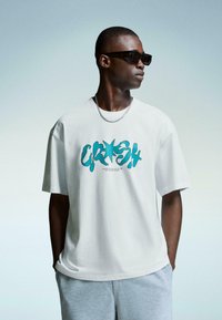 Bela bomba iz bombaža s turkizno grafiko, ki v valovnem pisanju pravi "CRASH", v kombinaciji s sivimi kratkimi hlačami in temnimi sončnimi očali.