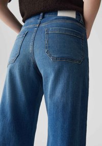 Blaue Denim-Jeans mit lockerer Passform, ausgestattet mit zwei Gesäßtaschen und einem weißen Lederpatch am Hosenbund. Weiche Textur, klassisches Design.