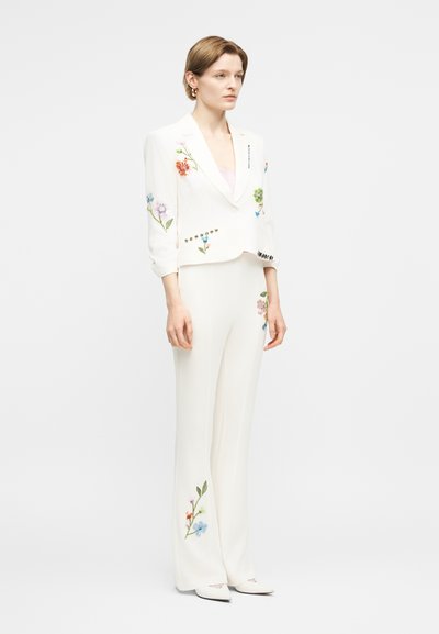 Γυναίκα που φοράει λευκή παντελόνα με πολύχρωμη κεντημένη ανθοφορία, στέκεται με τα χέρια χαλαρά μπροστά σε απλό λευκό φόντο.