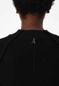 Maglione nero lavorato a maglia con un design a coste testurizzate e un piccolo dettaglio metallico sul retro del collo. Collo rotondo e cuciture sulle spalle.