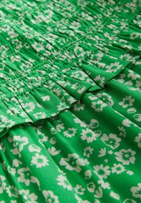 Zöld szövet fehér virágmintával, húzott textúrával és fodros díszítéssel a varrás mentén, rétegezett hatást keltve.