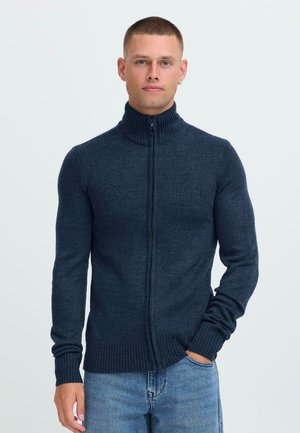 Mann mit kurzen Haaren, der einen dunkelblauen Reißverschluss-Pullover und hellblaue Jeans trägt. Er steht mit einer Hand in der Tasche vor einem schlichten Hintergrund.
