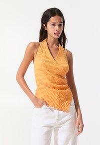 Oranžový halter top z texturované, zmačkané látky s hlubokým výstřihem do V, doplněný bílými kalhotami s vysokým pasem.