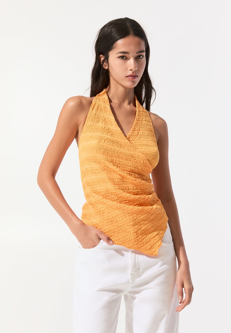 Oranžový halter top z texturované, zmačkané látky s hlubokým výstřihem do V, doplněný bílými kalhotami s vysokým pasem.