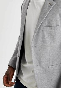 Blazer gris texturizado con solapa de muesca, un botón y bolsillo en el pecho, llevado sobre una camiseta gris clara.