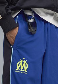 Blå atletiske shorts med sorte og lyseblå accenter, der har et fremtrædende gult logo og tekst på benet. Solbriller hviler på taljen.
