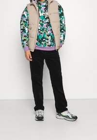 Chaleco acolchado beige sobre una sudadera con capucha de camuflaje verde con acentos morados, combinado con pantalones de pana negros y zapatillas grises con detalles verdes.