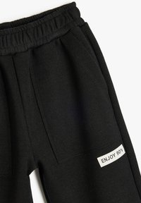Pantalones de chándal negros con cintura elástica, que cuentan con dos bolsillos frontales y una etiqueta blanca que dice "ENJOY 90's" en el lado izquierdo.