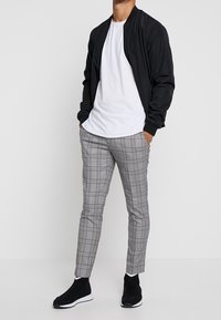 Czarna kurtka bomberka noszona na białej koszulce, zestawiona z szaro-czarnymi spodniami w kratę i czarnymi butami sportowymi z białą podeszwą.