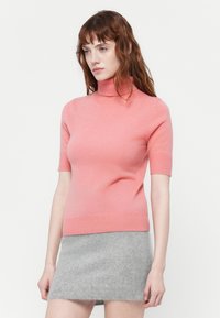 Jauna moteris su banguotais raudonais plaukais, vilkinti rožinį trumpą rankovę turtleneck megztinį ir pilką pūkuotą mini sijoną prieš baltą foną.