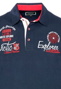 Marineblå polo skjorte med rød krave, præget med broderet tekst og grafik i hvidt og rødt. Bemærkelsesværdige detaljer inkluderer knapper og kontrasterende syninger.