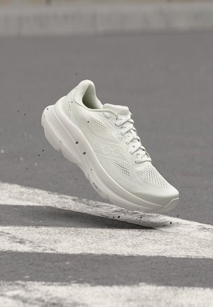 Chaussure de course blanche en suspension au-dessus d’une rue avec des lignes blanches de passage piéton et de petits débris près de la semelle.