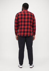 Camisa de cuadro roja y negra, de botones, de manga larga, vista posterior, combinada con pantalones oscuros y zapatillas blancas, mostrando un estilo casual.