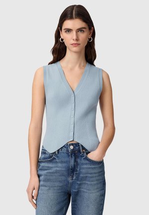 Kvinde iført en ærmeløs lyseblå ribbet vest med knapper og blå jeans, stående med den ene hånd i lommen foran en ensfarvet baggrund.