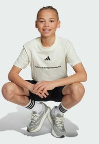 Adidas-Sport T-Shirt in Off-White mit schwarzem Logo, schwarze Shorts und graue Turnschuhe mit schwarz-weißen gestreiften Socken, in geduckter Position.