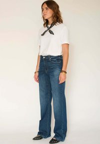 Jean en denim bleu taille haute avec jambes larges, associé à un t-shirt blanc et un bandana noir. Chaussures noires à bout pointu avec accents œillets.