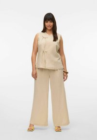 Haut beige sans manches avec liens à l'avant, associé à un pantalon ample assorti. Tissu au toucher doux, design minimaliste et chaussures jaunes.
