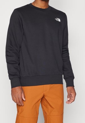 Homme portant un sweat-shirt noir à manches longues The North Face et un pantalon orange, debout devant un fond clair uni.