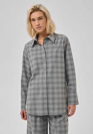 Persoon met schouderlang blond haar, gekleed in een loszittend grijs geruit overhemd met knopen en bijpassende broek, staand tegen een effen achtergrond.