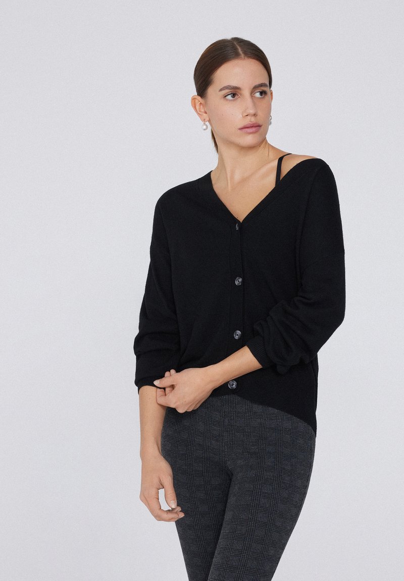 Schwarzer Cardigan mit V-Ausschnitt, langen Ärmeln und vorderem Knopfverschluss. Über einem dunkel gemusterten Outfit getragen, das einen weichen, strukturierten Stoff präsentiert.