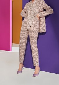 Světle barevný vzorovaný oblek s detaily vázanky, doplněný levandulovými lodičkami na vysokém podpatku. Pozadí tvoří barevné geometrické panely.