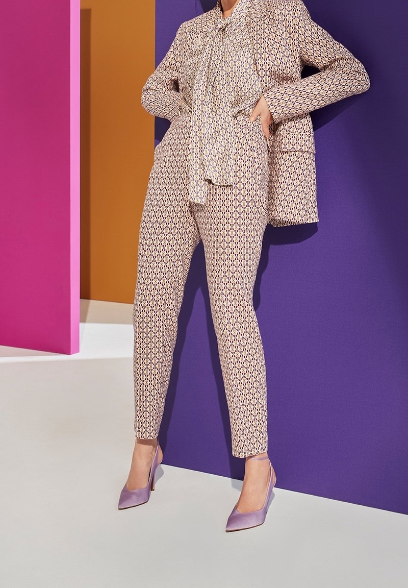 Světle barevný vzorovaný oblek s detaily vázanky, doplněný levandulovými lodičkami na vysokém podpatku. Pozadí tvoří barevné geometrické panely.