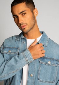 Giacca in denim azzurro chiaro con tasche sul petto con bottoni, cuciture a contrasto e una vestibilità rilassata, indossata sopra una camicia bianca.