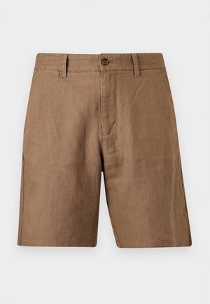Mann steht mit Händen in den Taschen, trägt ein gestreiftes Kurzarmhemd, braune Shorts, schwarzen Gürtel und schwarze Loafers vor einfarbigem Hintergrund.