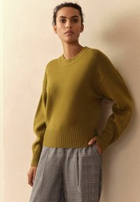 Frau trägt einen olivgrünen Strickpullover und graue karierte weit geschnittene Hosen, steht mit einer Hand in der Tasche vor einer beigen Wand.