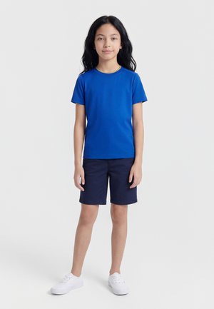 Mlada deklica z dolgimi črnimi lasmi stoji oblečena v modro kratko majico, temno modre kratke hlače in bele superge, na preprostem ozadju.