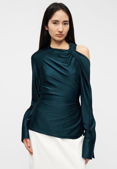 Γυναίκα με μακριά σκοτεινά μαλλιά, φορώντας ένα ασύμμετρο τοπ από σατέν σε τιρκουάζ χρώμα με έναν ώμο ακάλυπτο και μακριά μανίκια, σε συνδυασμό με λευκή φούστα.