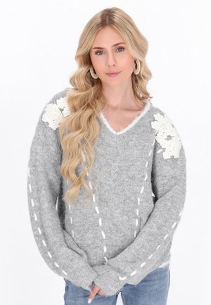 Blonde vrouw met lang gekruld haar draagt een grijze gebreide trui met witte bloemmotieven op de schouders en witte gestikte lijnen op de mouwen en voorkant.