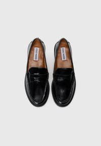 Par med svarte skinn slip-on loafers med rund tå og synlig Steve Madden-merke inne i hver sko mot en hvit bakgrunn.