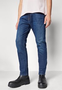 Jeans de mezclilla azul oscuro con corte ajustado, que presentan una textura sutil y un diseño clásico de cinco bolsillos, combinados con botas negras hasta el tobillo.